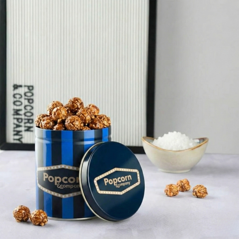 
                  
                    Blue and white striped popcorn container with 'Popcorn Company' branding, filled with popcorn, on a light gray surface.
                  
                